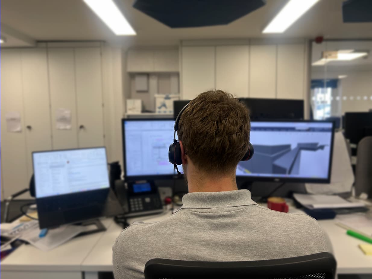 A person wearing headphones sits at a desk with three monitors in front of them, working in an office environment. The screens display emails and design software.