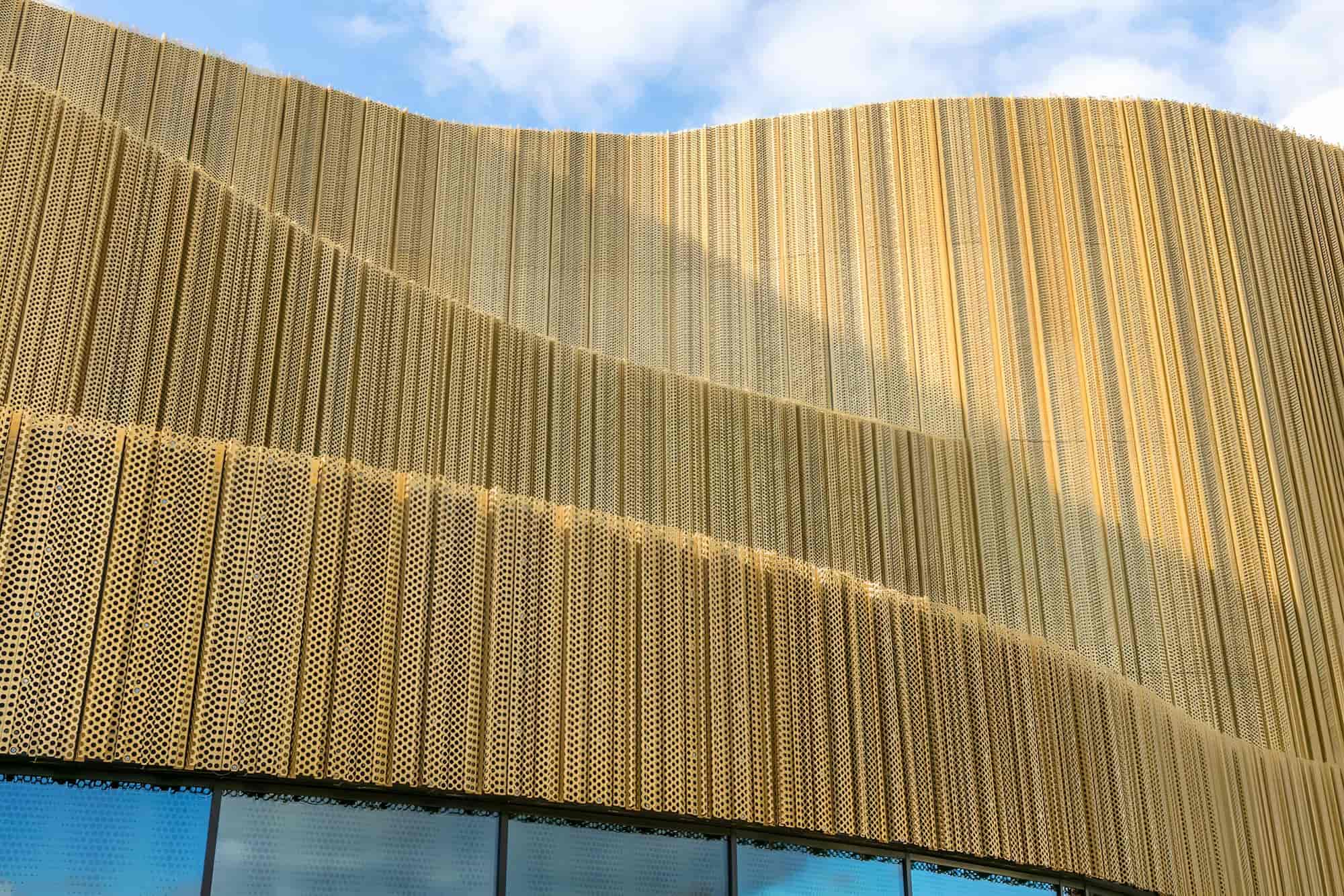 Curved golden metal facade with perforated panels on a modern building against a partly cloudy blue sky.