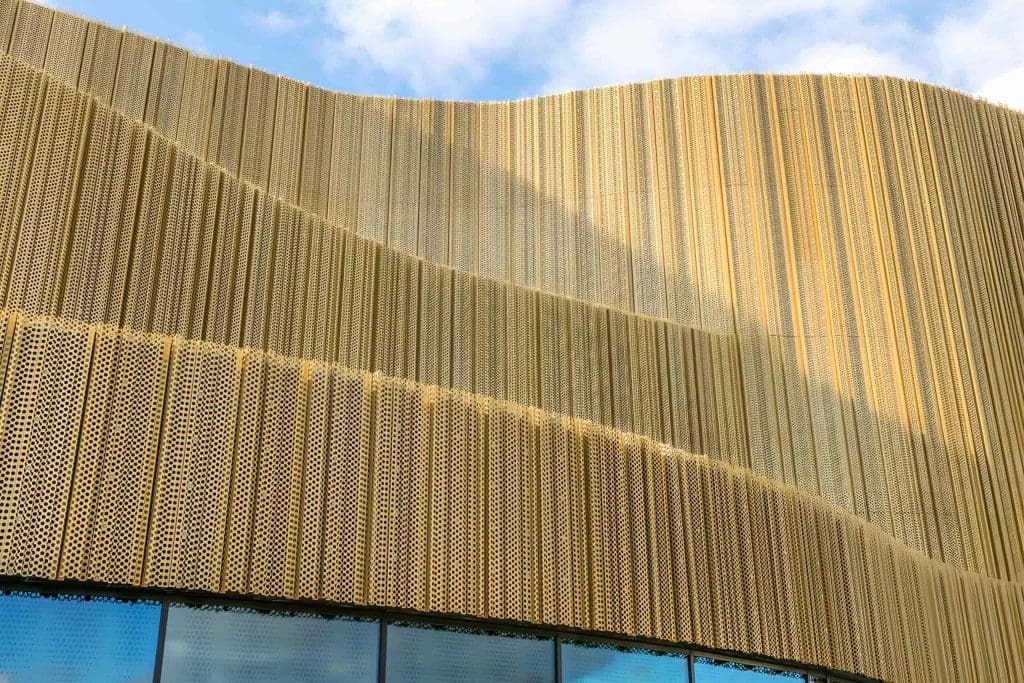 A modern building facade with wavy, gold perforated metal panels and a row of reflective glass windows at the bottom, set against a blue sky with clouds.