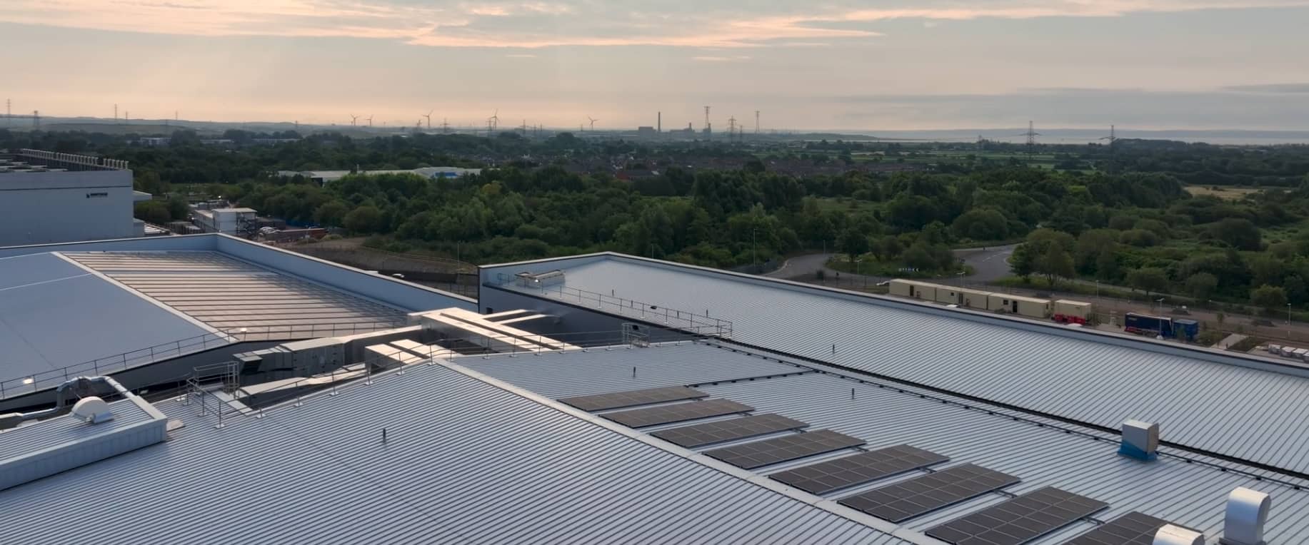 A large industrial building with solar panels on its metal roof, surrounded by trees, roads, and distant wind turbines under a cloudy sky at sunset.