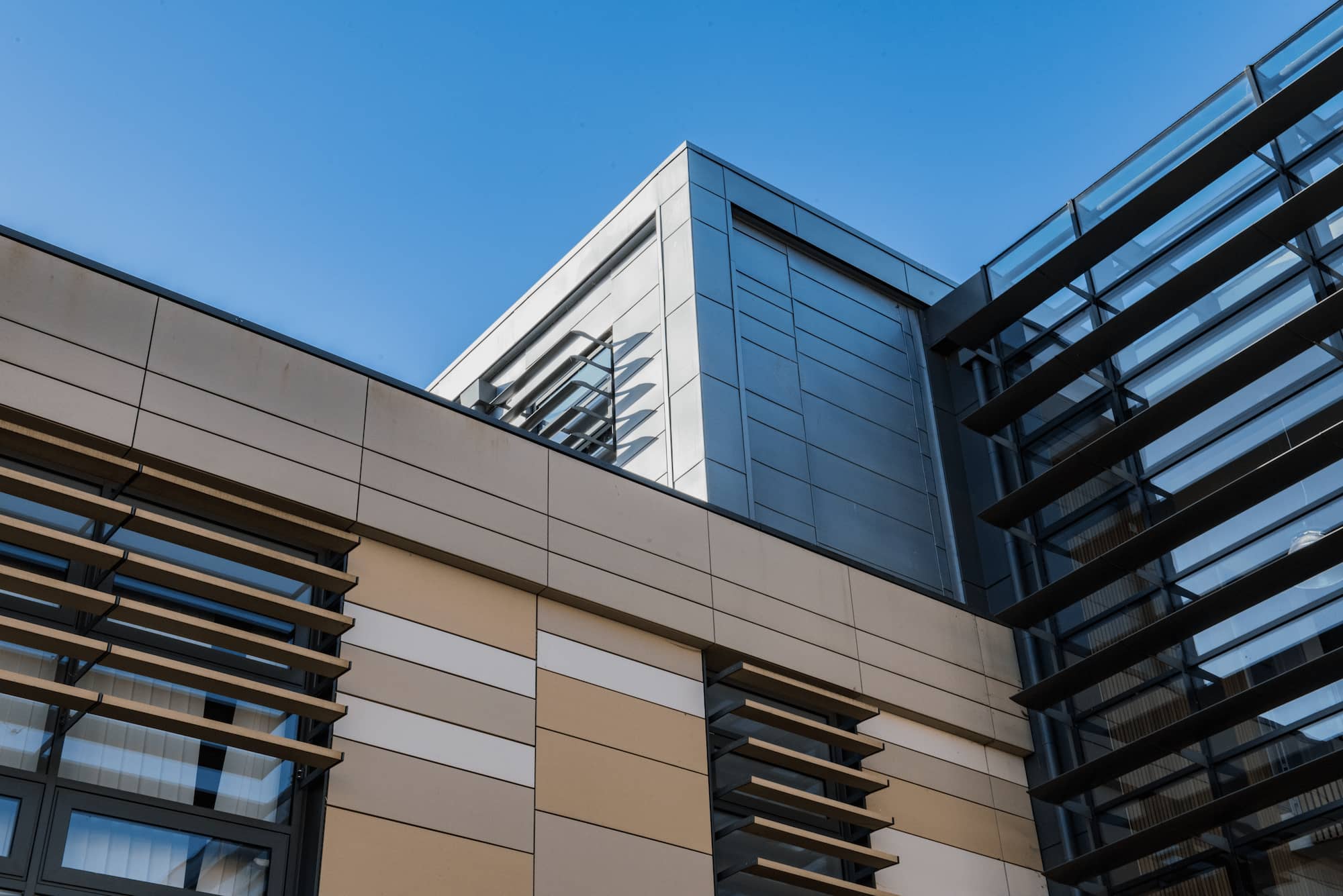Modern building exterior with tan and cream panels, large windows, horizontal metal sunshades, and a section of reflective glass under a clear blue sky. The architecture features sharp angles and clean lines.