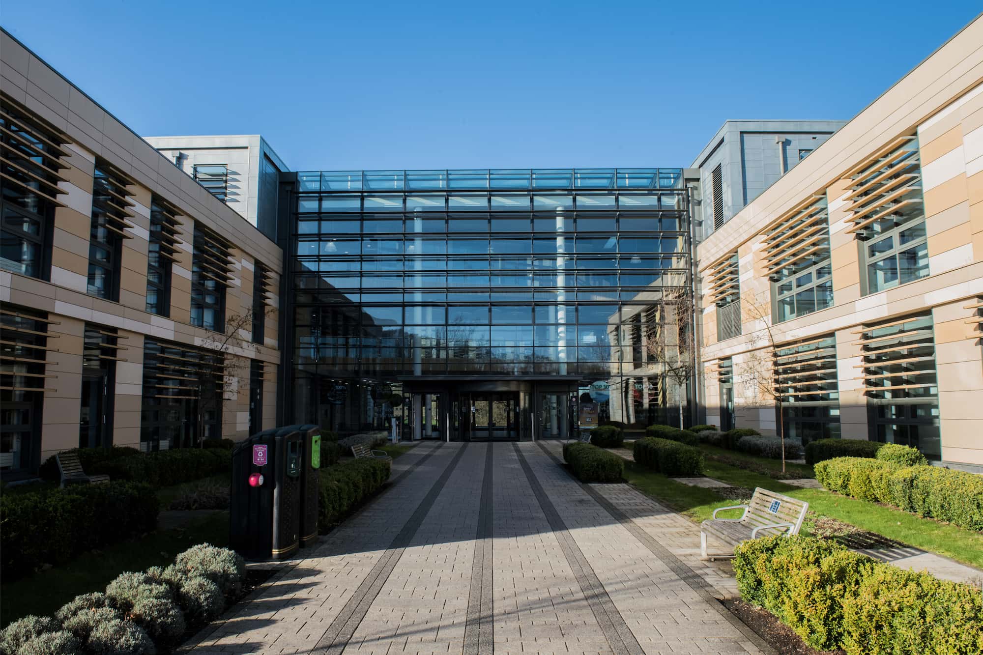 Modern glass-fronted building with two wings, landscaped bushes, benches, and a clear blue sky. A paved pathway leads to the main entrance at the center of the image.