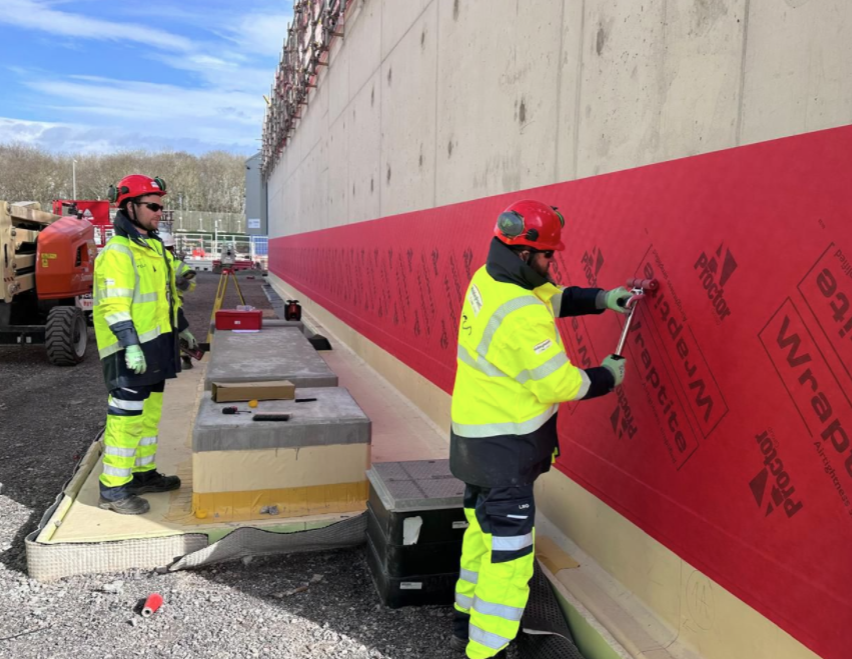 Two construction workers in high-visibility jackets and red helmets apply red waterproofing wrap to a concrete wall at a construction site. Equipment and materials are nearby. Trees are visible in the background.