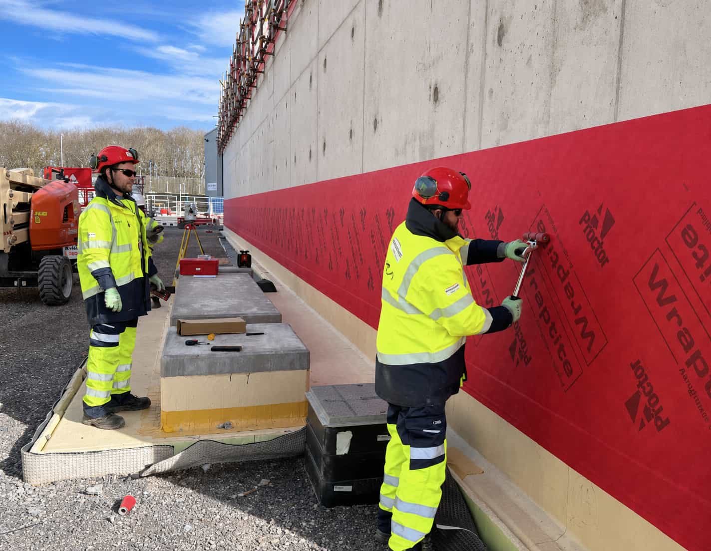 Two construction workers in high-visibility jackets and helmets apply red waterproofing membrane to a concrete wall at an outdoor construction site under a blue sky.